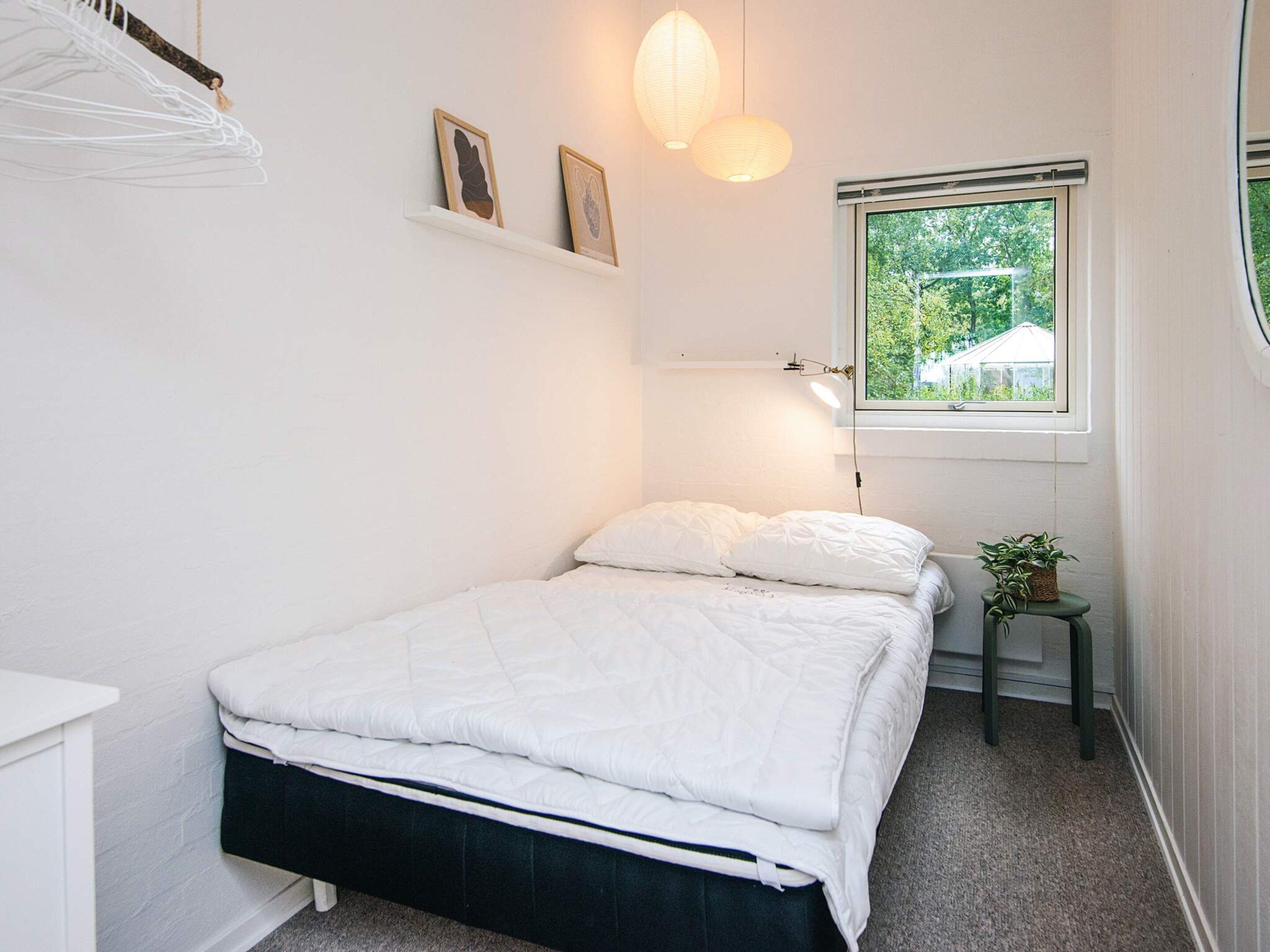 Un dormitorio con cama, mesita de noche con lámpara, ventana con vista a follaje y estantes con marcos que albergan fotografías.