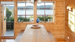 Una cabaña de madera con una gran mesa de comedor, ventanas amplias con vistas a un lago y montañas, y una terraza de madera exterior.