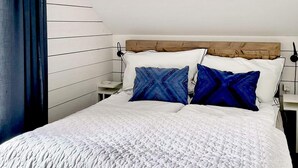 Una cama bien tendida con colcha blanca y dos almohadas azules, un cabecero de madera y una mesita de noche con lámpara.