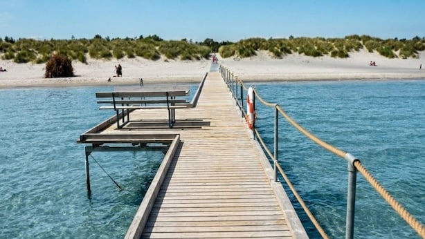 A wooden pier extending into clear blue water with a sandy beach and dunes in the background. - 4 Star Holiday Home in Jerup (Jerup)