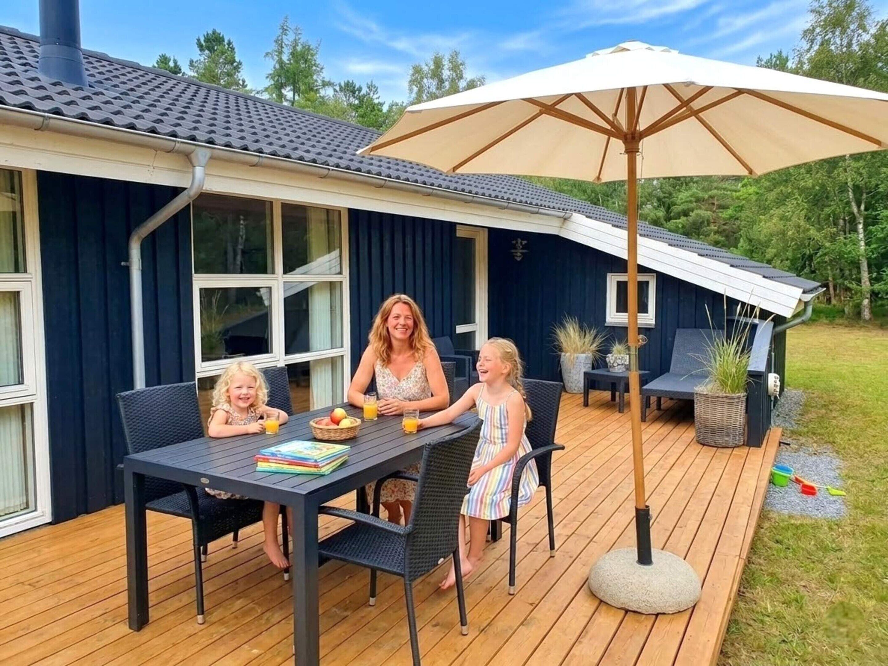 Een gezin dineert aan een buitentafel met een grote parasol, een houten terras en een huis met donkere bekleding.