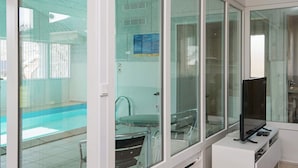 A modern hotel room with large glass doors leading to a pool area, a flat-screen TV, and a small seating area with a table and chairs.