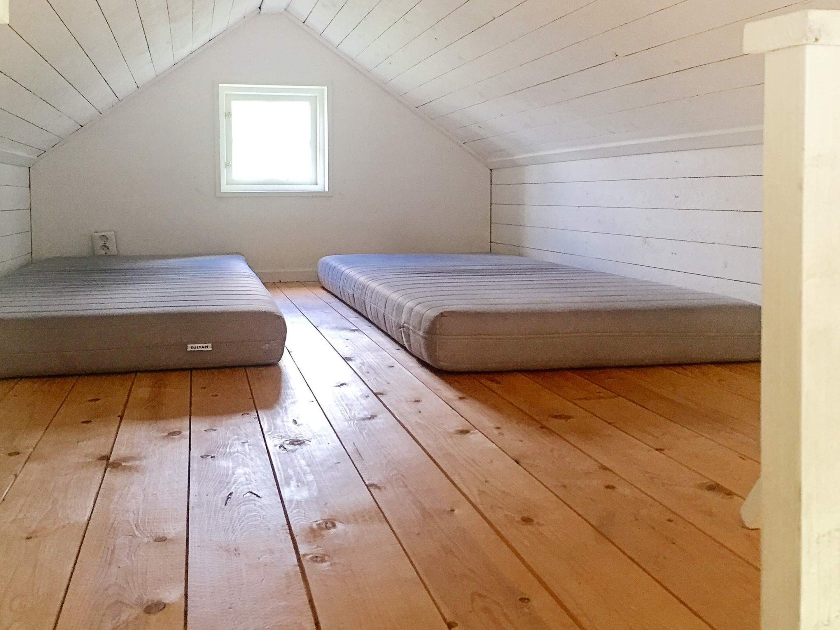 A small attic room with wooden floors, two mattresses, a window, and a white wall.
