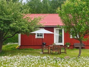 A red wooden house with a white door, a small porch with a table and chairs, and a garden with white flowers. - 4 Person Holiday Home in Stenhamra (Stenhamra)