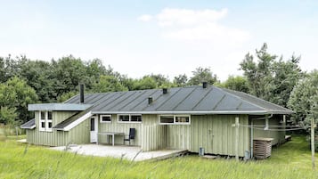 A single-story wooden house with a sloped roof, a porch with a bench, and a small outdoor table.