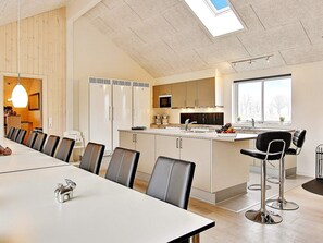 Eine moderne Küche mit einer zentralen Kochinsel, einem Essbereich mit langem Tisch und Stühlen sowie einer Aussicht ins Freie durch ein großes Fenster.