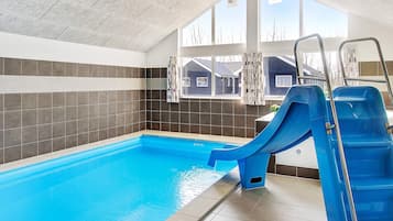 An indoor swimming pool with a blue slide, tiled walls, and a view of a residential area through large windows.