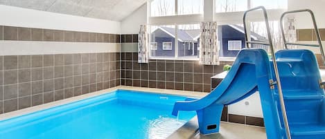 En inomhuspool med en blå vattenrutschkana, kaklade väggar och utsikt över ett bostadsområde genom stora fönster.