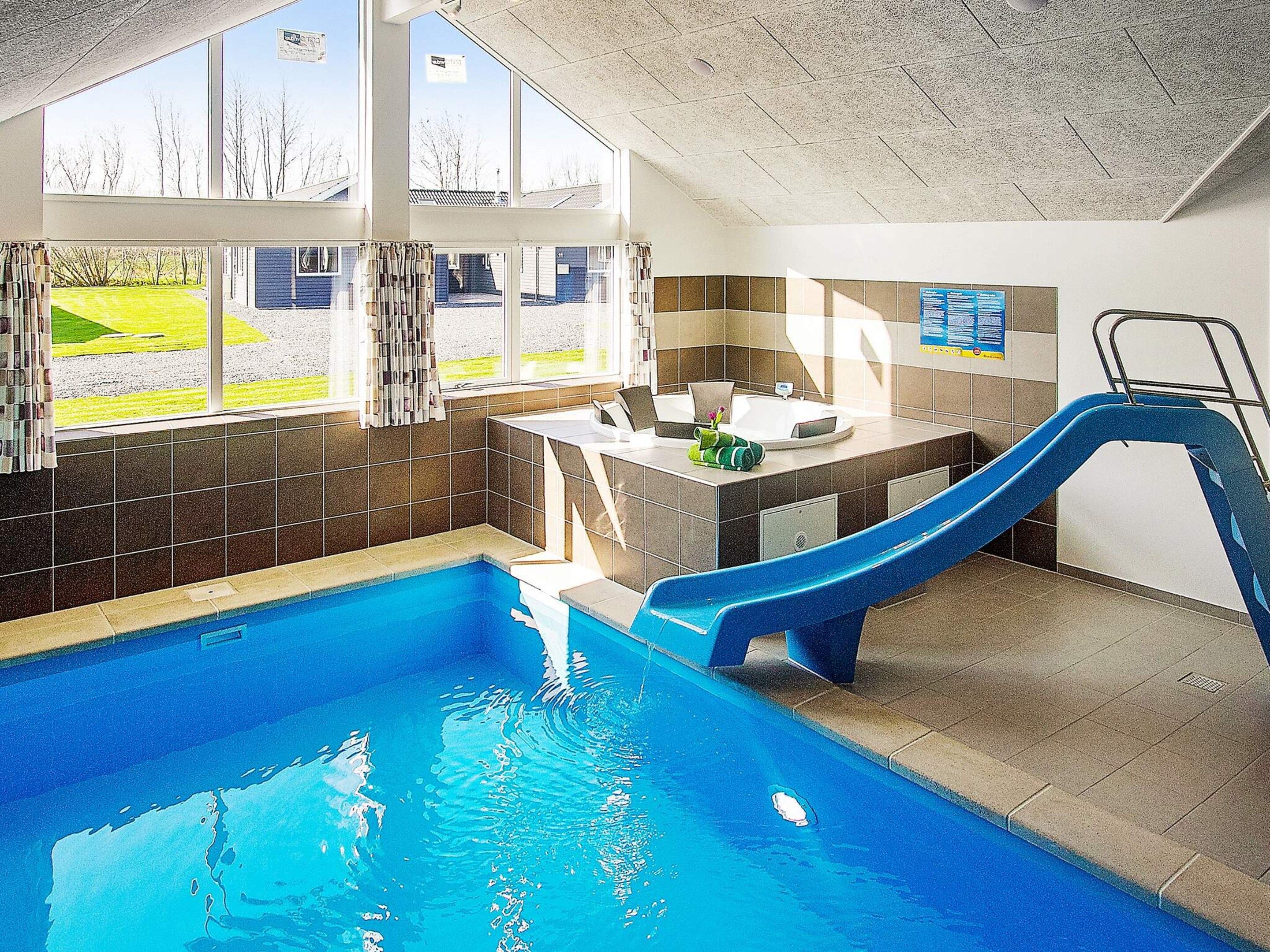 An indoor swimming pool with a blue slide, a changing room, and a view of a grassy area through large windows.