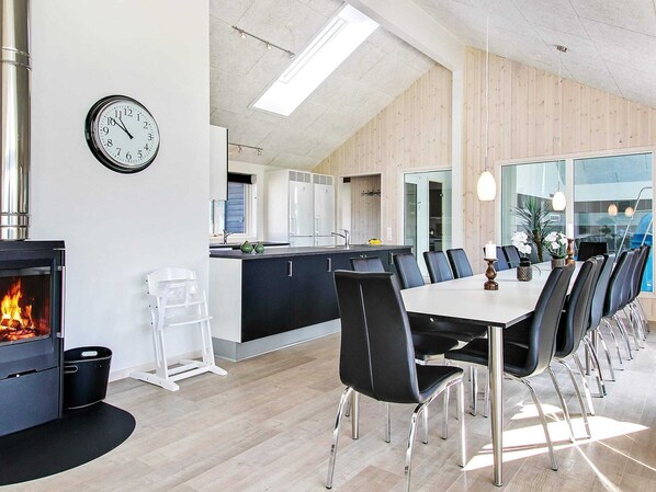 A modern kitchen with a central island, a dining area with a long table and chairs, a visible clock, and a fireplace.