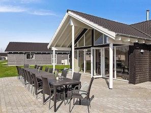 Ein überdachter Essbereich im Freien mit einem langen Tisch und Stühlen, einer Pergola und einem Gebäude mit großen Fenstern und einem geneigten Dach.