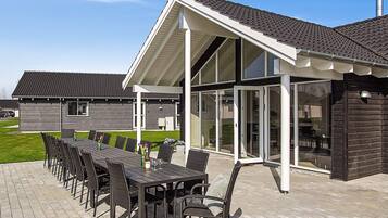 A covered outdoor dining area with a long table and chairs, a pergola structure, and a view of a residential building in the background.