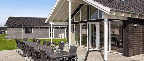 Un área de comedor al aire libre techada con una mesa larga y sillas, una estructura de pérgola y vista a un edificio residencial al fondo.