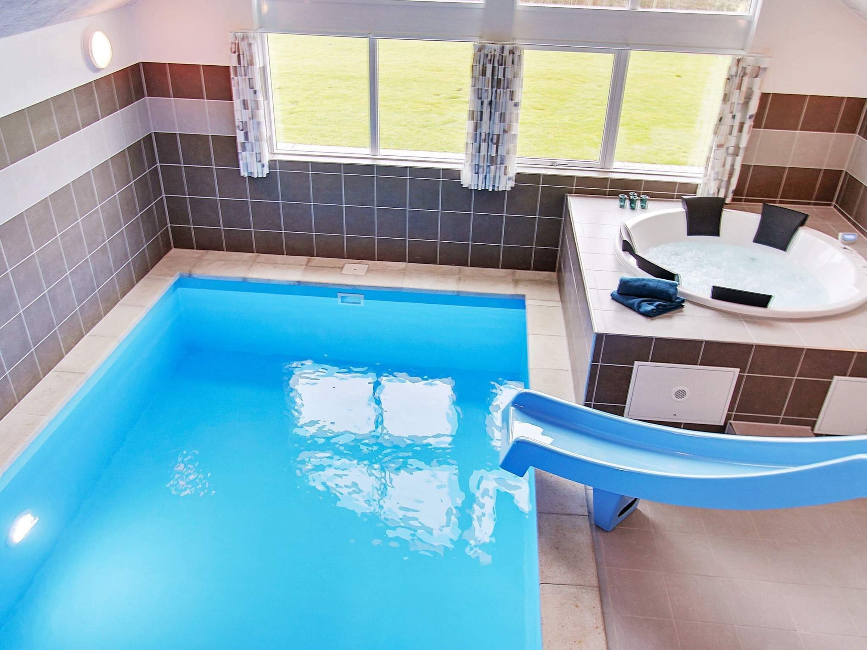 Une piscine rectangulaire avec un toboggan bleu, un jacuzzi et une vue sur une zone verdoyante, visible à travers une fenêtre.