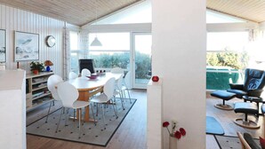 A modern dining area with a wooden table and white chairs, a large window with a view of greenery, and a black chair in the corner. - 4 Star Holiday Home in Hadsund (Hadsund)