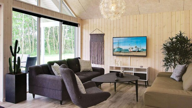 Un salon moderne avec un canapé, un fauteuil et une table basse, de grandes fenêtres et un téléviseur fixé au mur.
