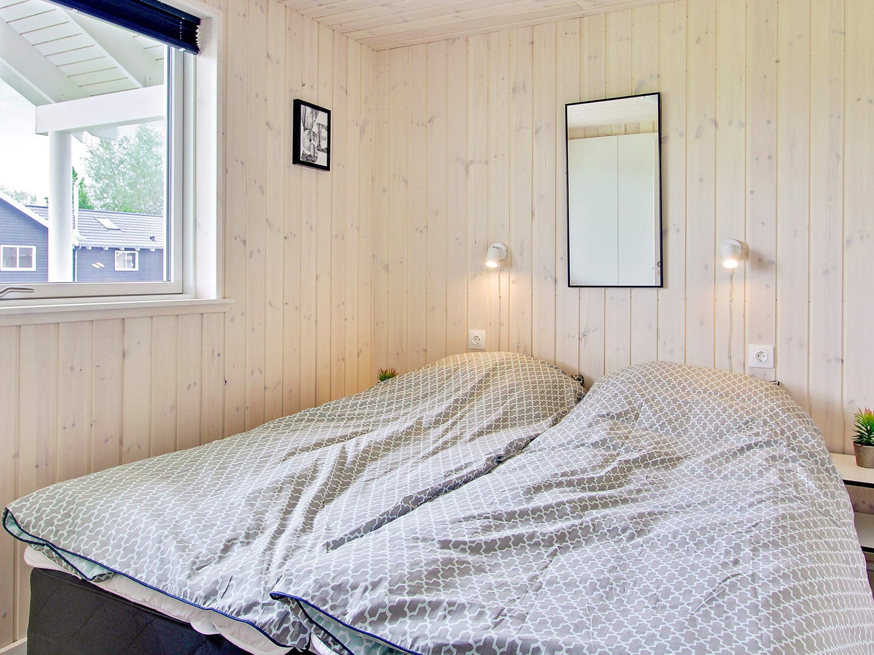 A room with wooden panel walls, a bed with patterned bedding, a window with blinds, and wall-mounted lights.