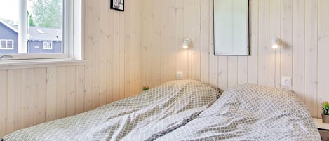 A room with wooden panel walls, a bed with patterned bedding, a window with blinds, and wall-mounted lights.