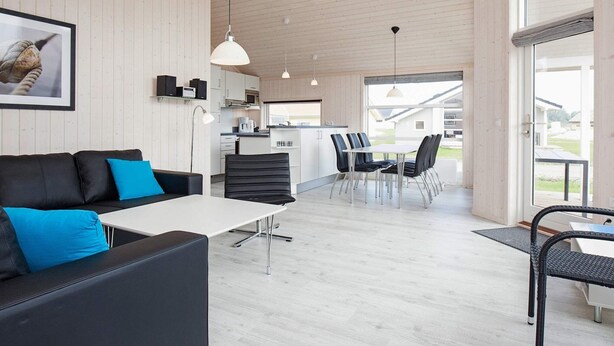 Un intérieur moderne avec un canapé en cuir noir, une table blanche et un coin repas avec des chaises. On aperçoit une cuisine au fond, avec vue sur l’extérieur.
