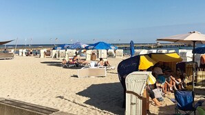 Une plage avec des transats et des parasols, une jetée en arrière-plan et des drapeaux sur des mâts.