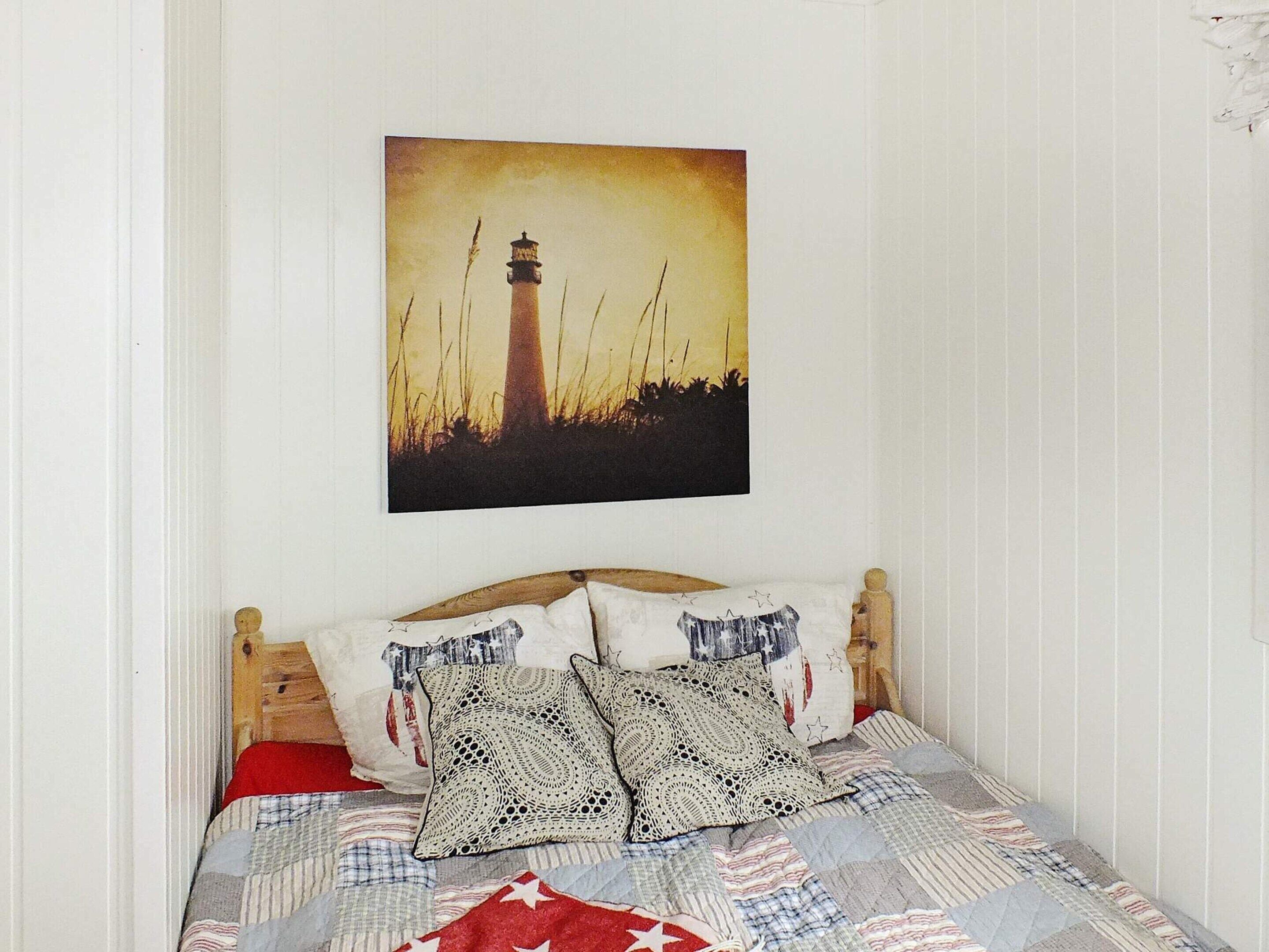 Una cama con colcha, dos almohadas estampadas y un cuadro en la pared de un faro.