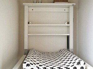 A small, narrow room with a white bed and a headboard with shelves.