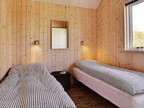 A room with wooden panel walls, two beds with striped bedding, a small black ottoman, and a window with a view of greenery.