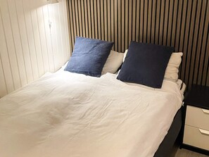 Una cama con ropa blanca y dos almohadas, una mesita de noche con cajón, y una pared con listones de madera verticales.