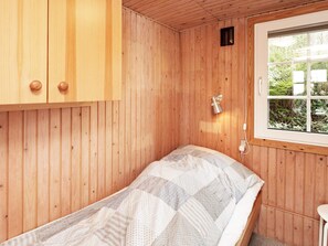 A wooden room with a bed, a window with a view of greenery, and a wall-mounted lamp.