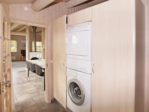 A laundry room with a washing machine and dryer, wooden cabinets, a dining area with a table and chairs, and a view through an open door to another room.