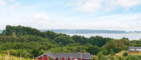 Une maison rouge avec un toit en pente, entourée d’arbres verdoyants et avec une étendue d’eau en arrière-plan.