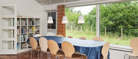 A dining area with a long table, chairs, a bookshelf, and a view of greenery outside.
