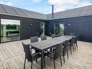 A modern outdoor dining area with a long table and chairs, a chimney, and a view through large glass doors.