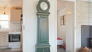 A room with a large green clock, a kitchen area with a white oven, and a door leading to another room with a bed and a fireplace. - 6 Person Holiday Home in Hasle-by Traum (Hasle)