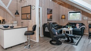 Una habitación tipo loft acogedora, con techos altos, paredes de madera, zona de comedor con barra, un sofá de cuero y una mesita con lámpara.