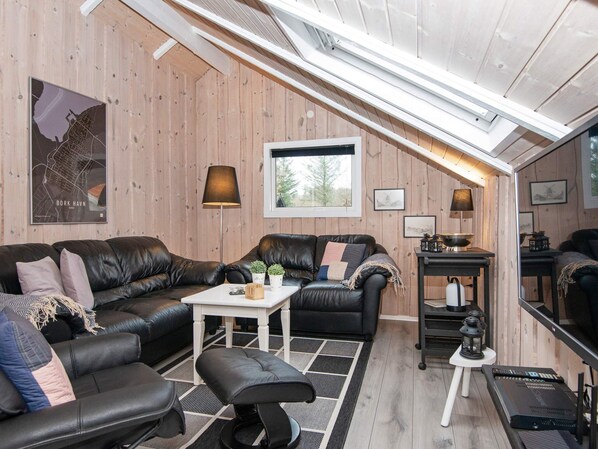 A cozy loft-style living room with a high ceiling, wooden walls, a large window, and a black leather sofa set.