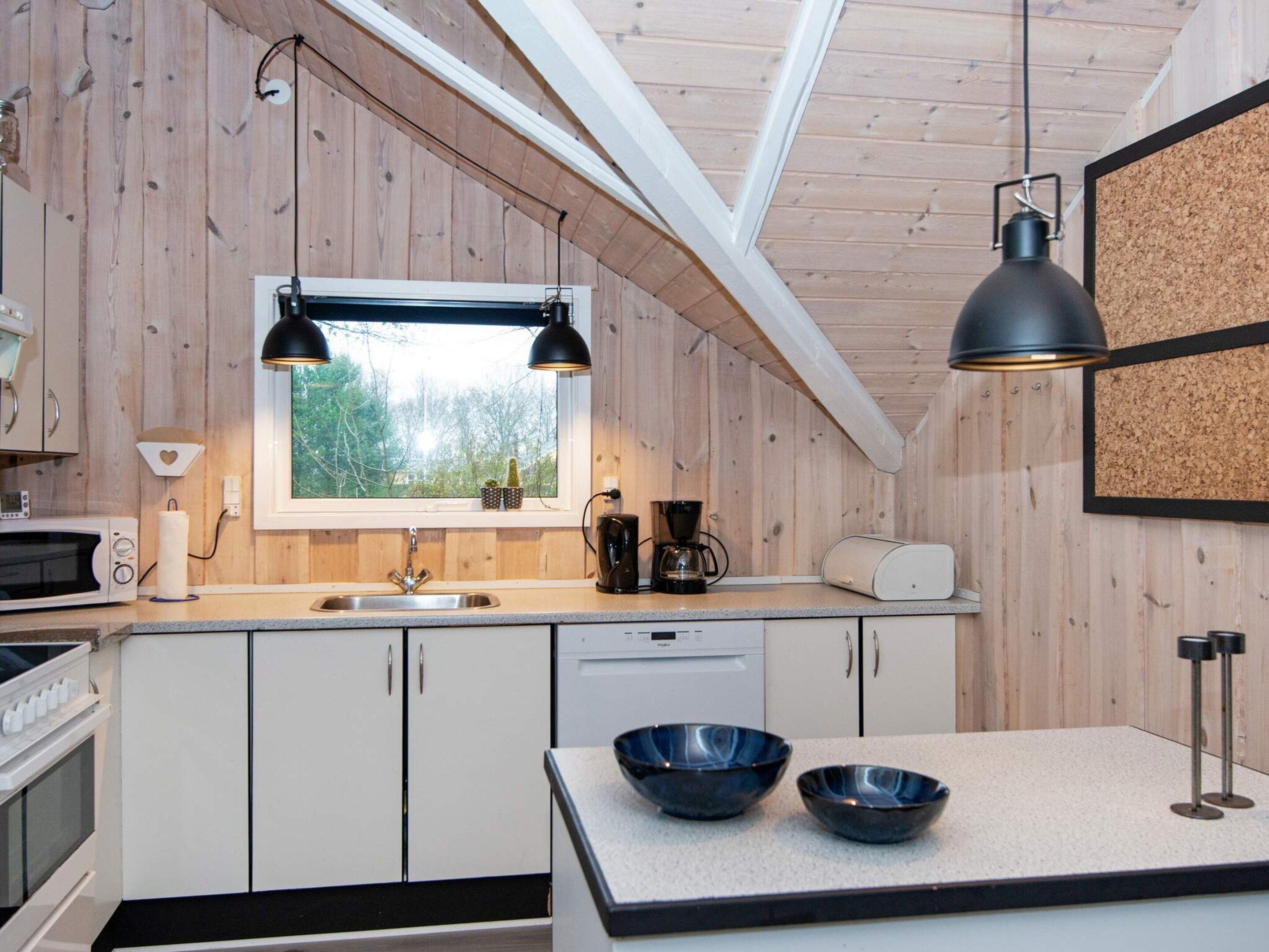 A compact kitchen with wooden walls, a window, a sink, a microwave, a toaster, and two bowls on the counter.