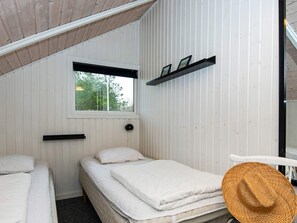 A small, single-room accommodation with a sloped ceiling, a window with a view of greenery, a bed with a white mattress and pillows, a bedside table with a lamp, and a straw hat on a chair. - 6 Person Holiday Home in Hemmet (Hemmet)