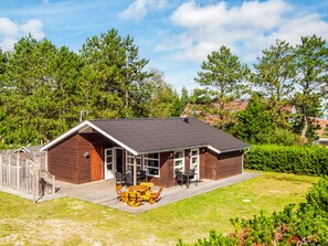 A wooden cabin with a deck, a patio with outdoor furniture, and a view of a forested area.