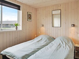 A room with wooden panel walls, a bed with patterned bedding, a window with a view of a building, and a framed picture.