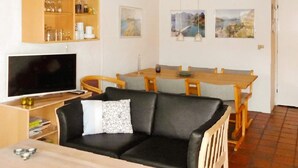A living room with a black leather sofa, a wooden dining table with chairs, a television, and a bookshelf with various items. - Cozy Beach House in Blavand - By Traum Ferienwohnungen (Blavand)