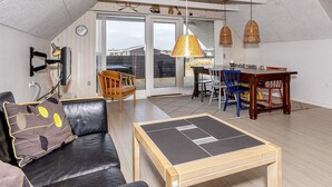 A modern loft apartment with a sloped ceiling, a dining area with a wooden table and chairs, a leather sofa with a patterned cushion, and a large sliding glass door leading to a patio. - 6 Person Holiday Home in Thisted-by Traum (Thisted)