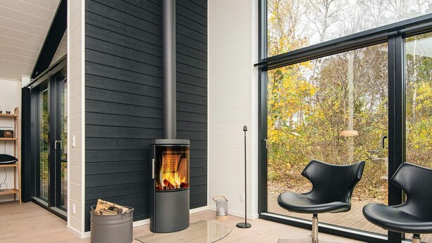 A modern living room with a black wood-burning stove, a large window with a view of trees, and a white table with glasses.