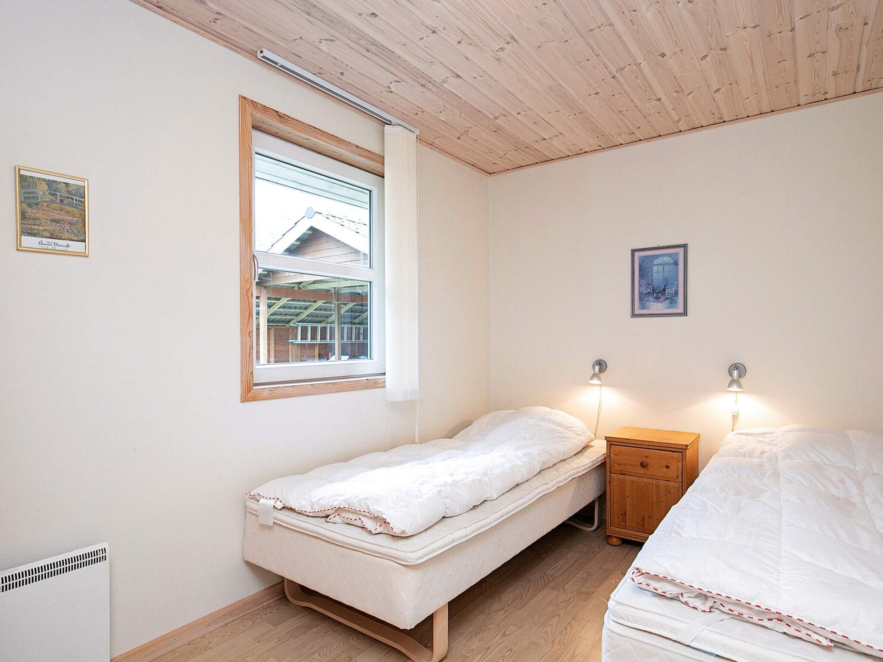 A small, single-bed room with a window, a bedside table, and a lamp.