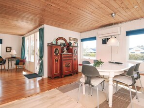 Un espace repas avec un plafond en bois, une table blanche, des chaises grises, un buffet en bois et une grande fenêtre.