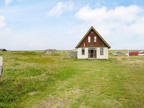 A small, single-story building with a triangular roof, situated in a grassy field with a few other structures in the background. - Scenic Holiday Home in Harboøre near Sea (Harboore)