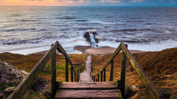 Un escalier en bois mène à une plage d’où l’on peut admirer l’océan et un ciel nuageux.
