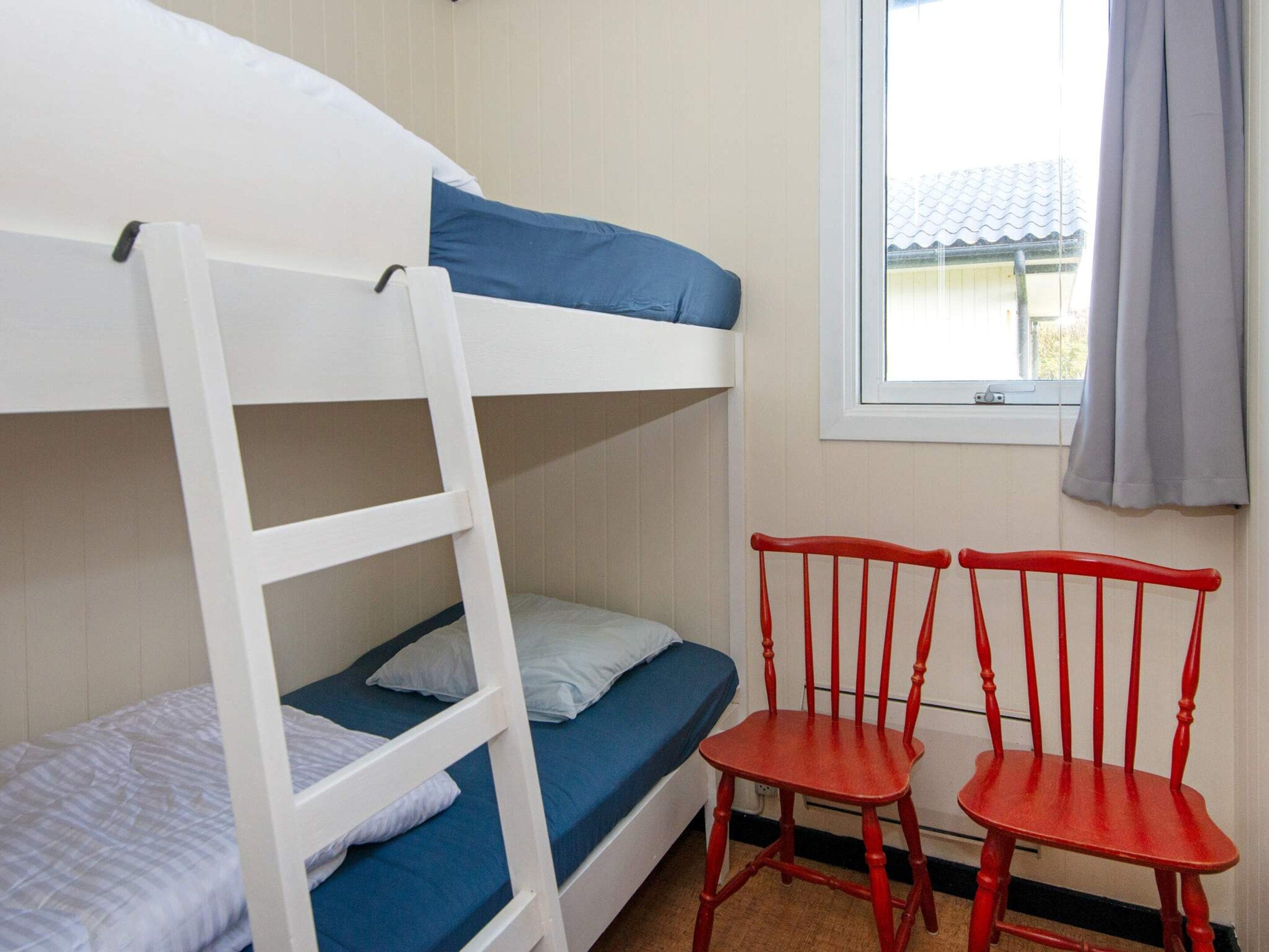 Una cama litera con escalera, dos sillas rojas y una ventana con vista a un edificio.