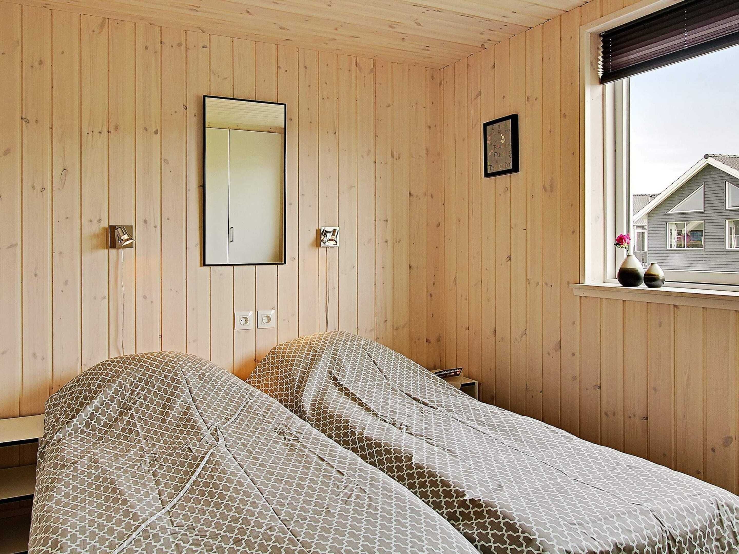 A room with wooden panel walls, a bed with patterned bedding, a mirror, and a window with a view of a house.