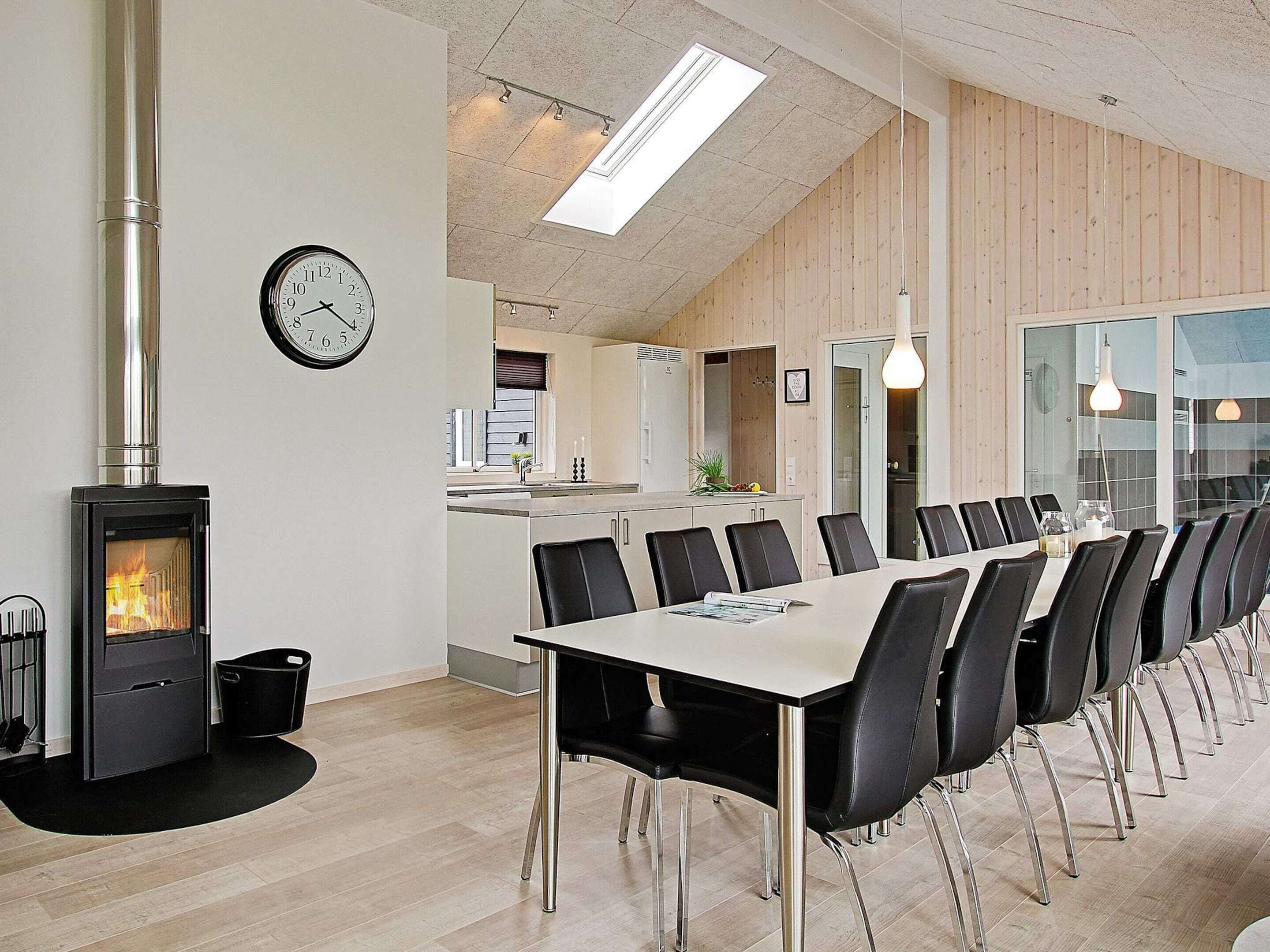 A modern dining area with a long table, chairs, a wood-burning stove, and a clock on the wall.